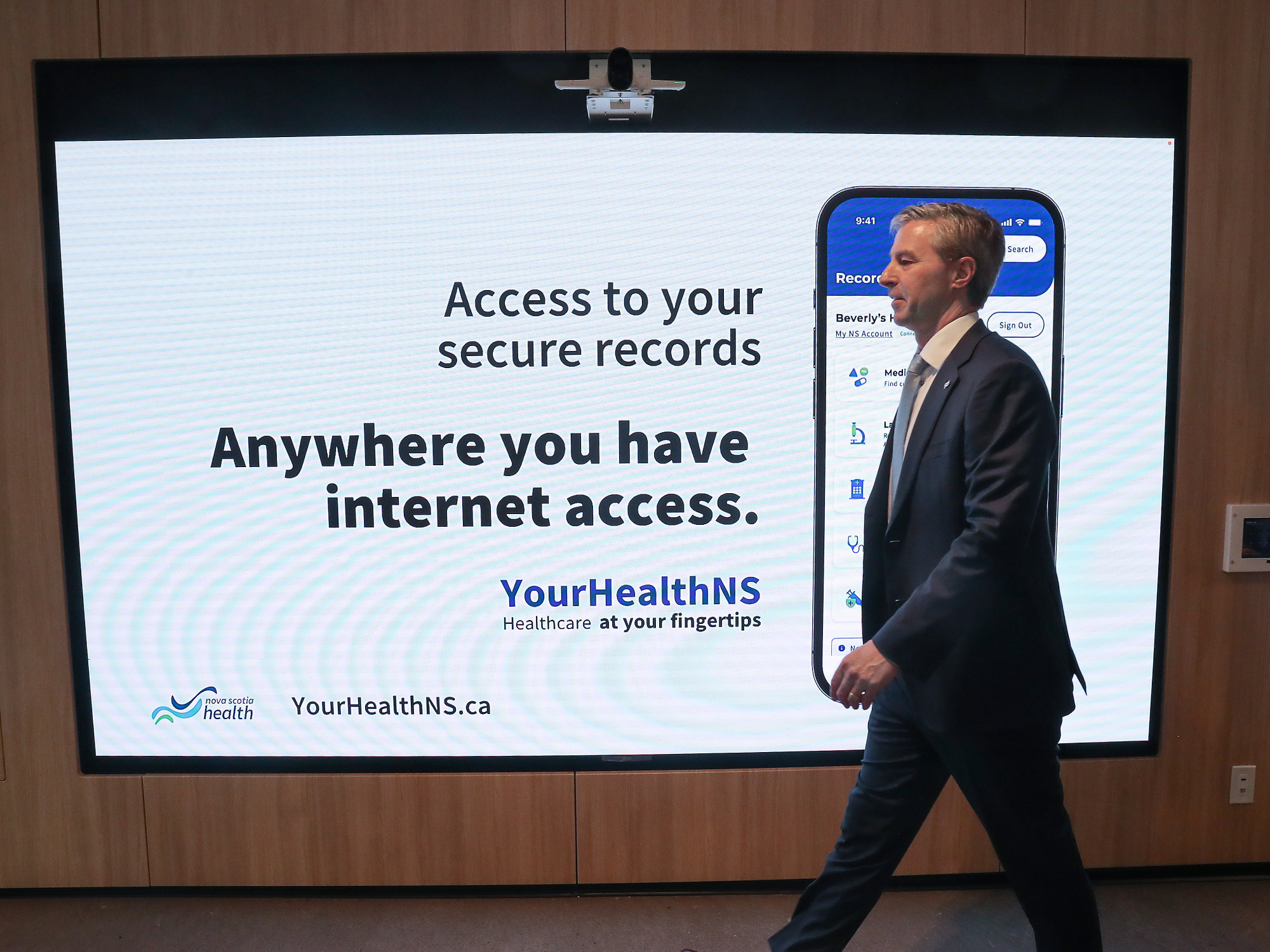 Photo of Tim Houston walking in front of a screen showing information about access to records on YourHealthNS