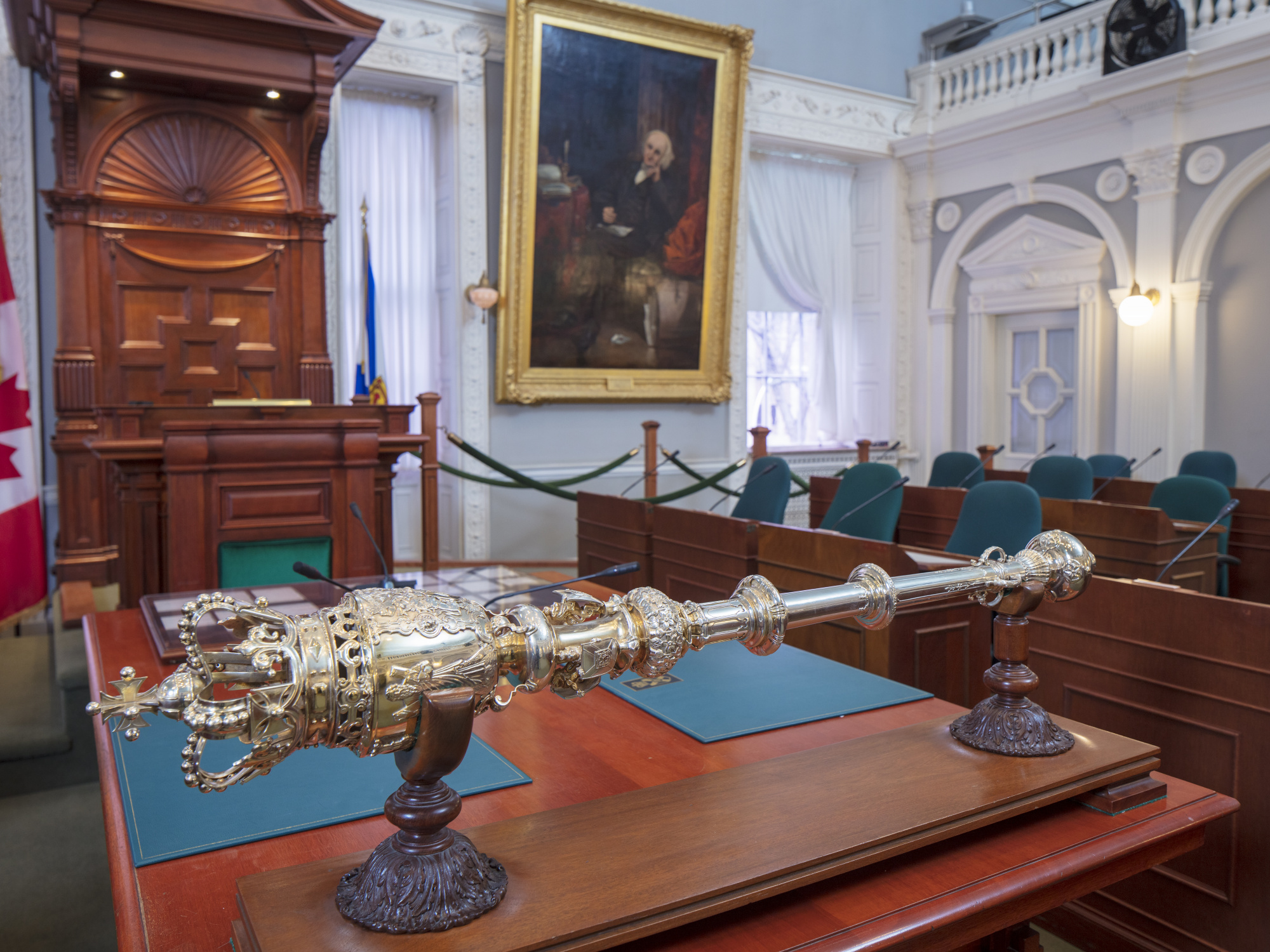 Photo showing mace sitting on a table in front of MLAs’ desks in the legislative chamber at Province House. The Speaker’s desk and a large portrait are in the background