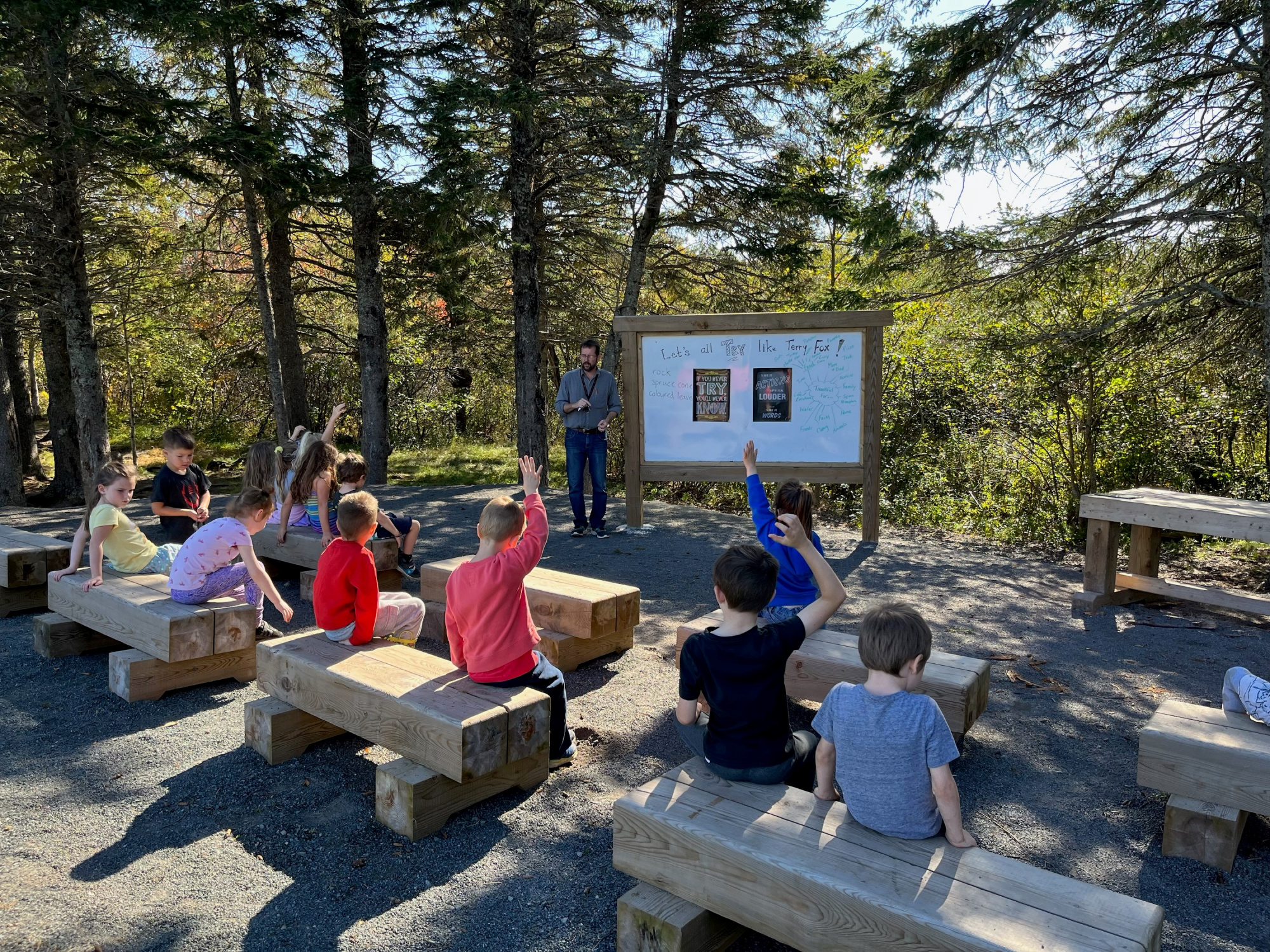 Photo of young students sitting on wooden benches surrounded by trees as their teacher speaks to them from beside an information board