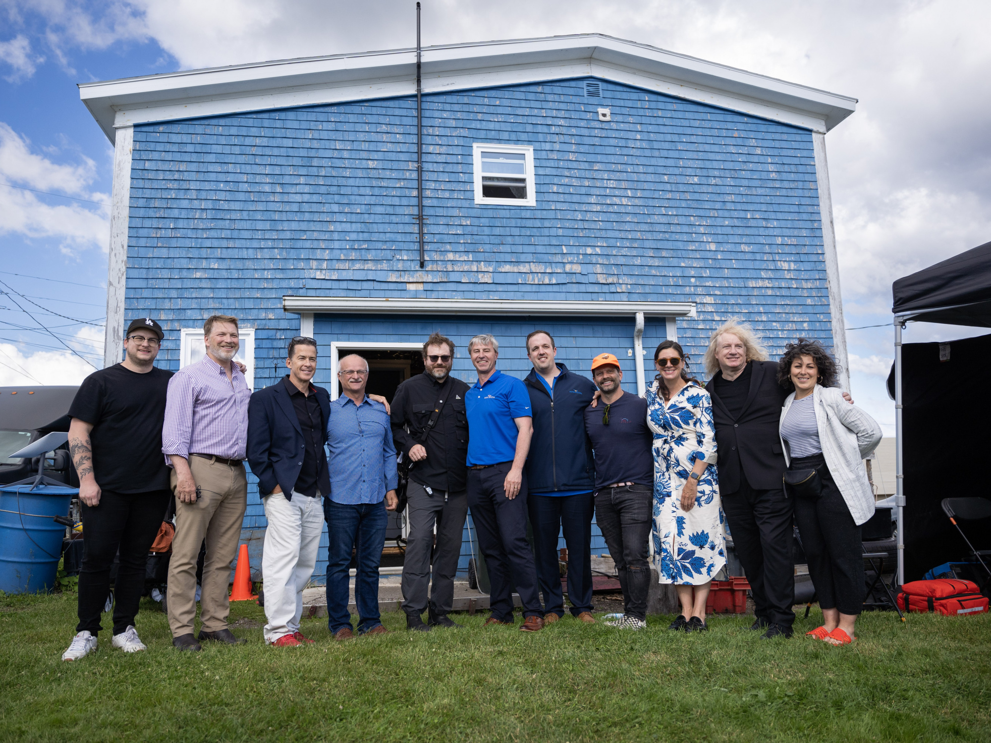 Photo of Premier Tim Houston with a group of people in front of a blue shingled building on the set of Little Lorraine in Louisbourg