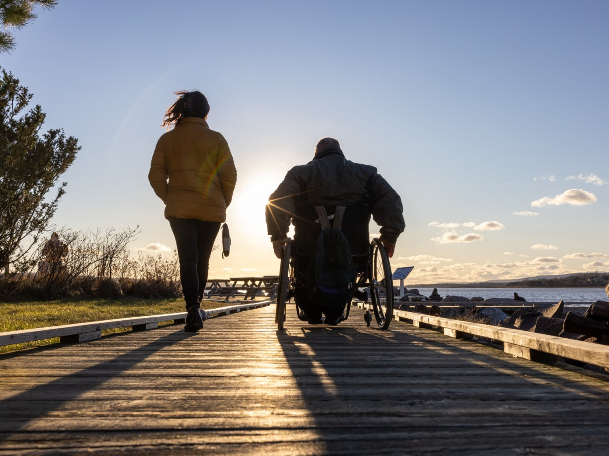 Person walking on a trail beside a person using a wheelchair, at sunrise.