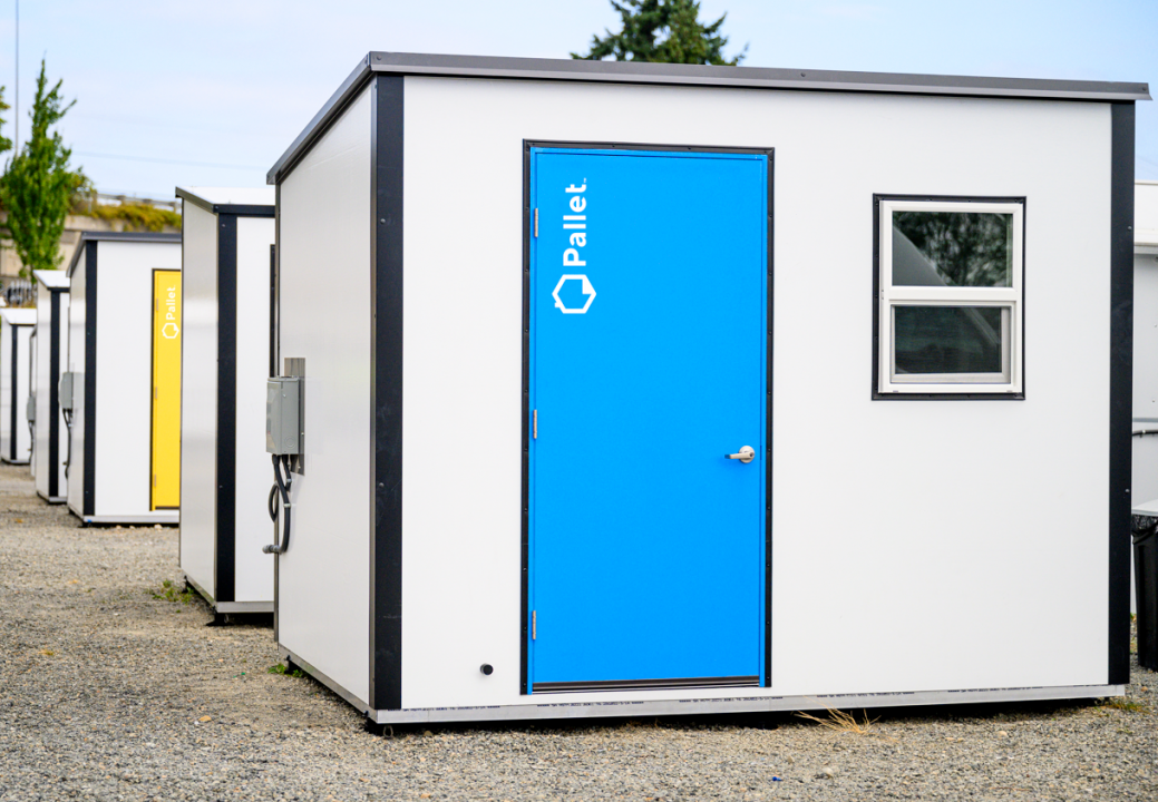 Photo showing a white Pallet shelter with a blue door, with other shelters in the background