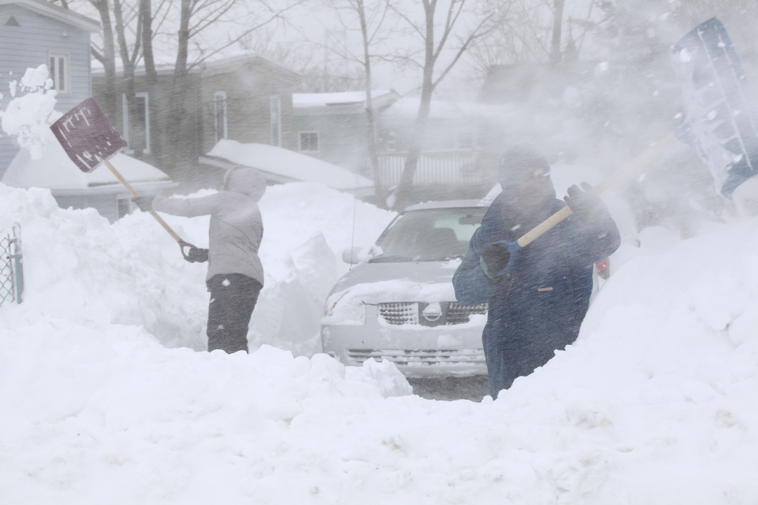 photo of two people shovelling a driveway as the snow blows around them
