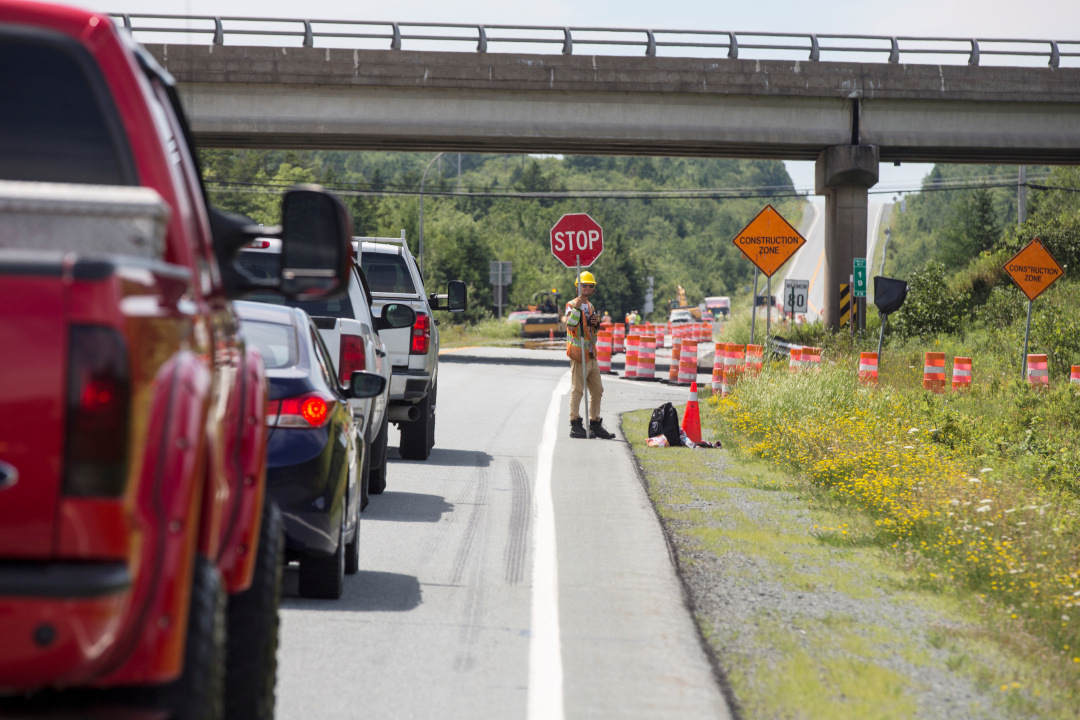 A photo showing a line of vehicles stopped on a highway behind a worker holding a sign that says Stop.