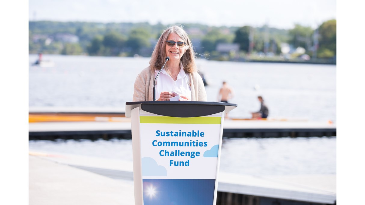 A woman stands behind a podium speaking with Lake Banook in the background