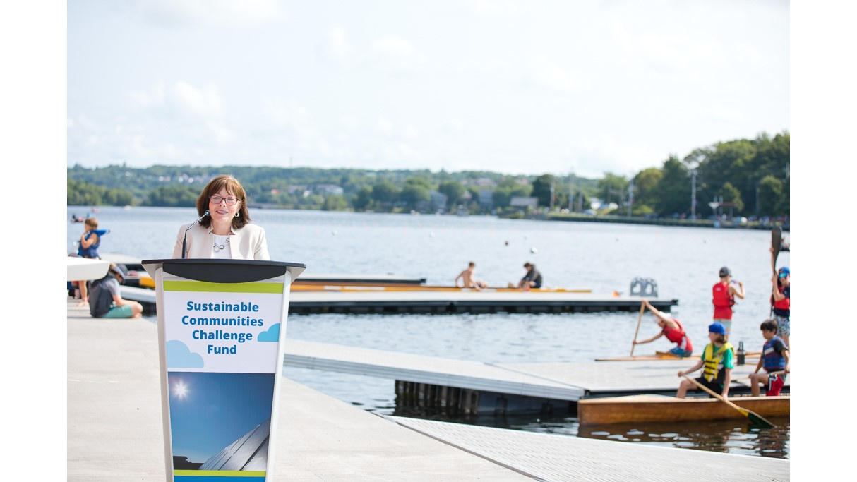 A woman speaks from a podium with a lake in the background.