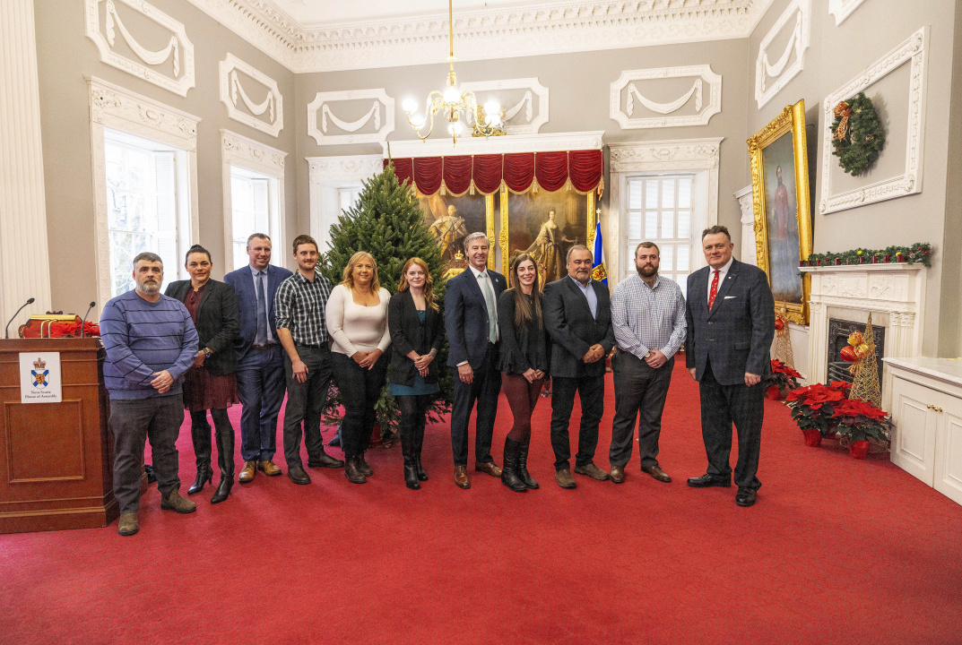 Photo of Premier Tim Houston, Lt.-Gov. Mike Savage and a group of people in front of an undecorated Christmas tree in the Red Chamber at Province House