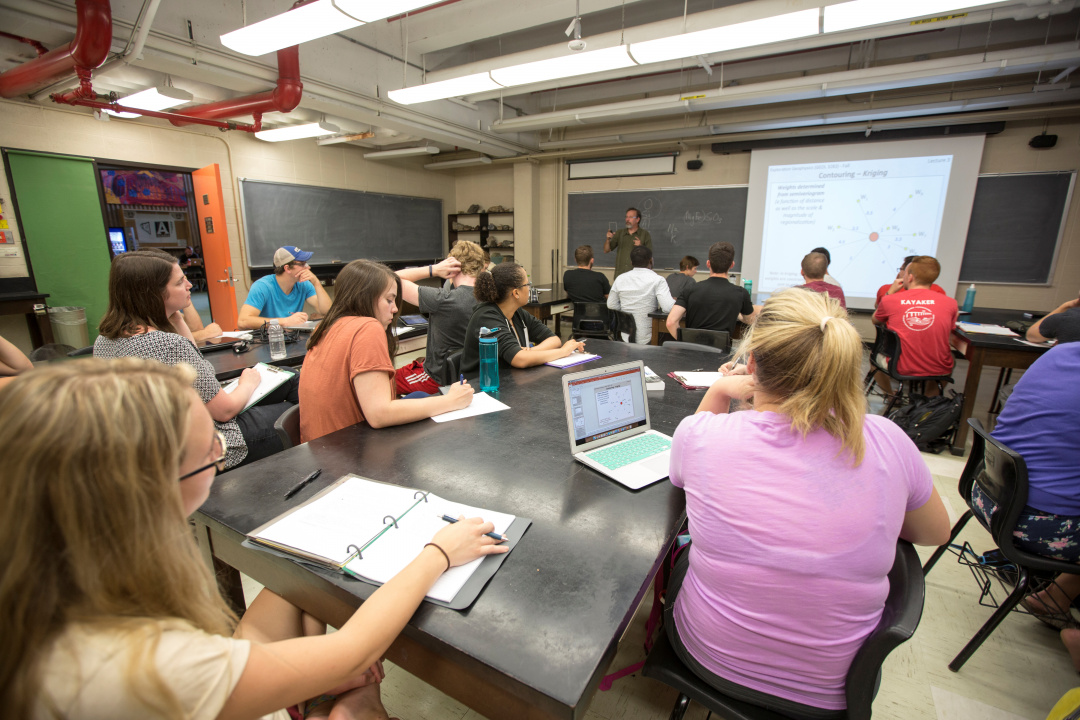 Students in a classroom sitting at tables looking towards a projector screen