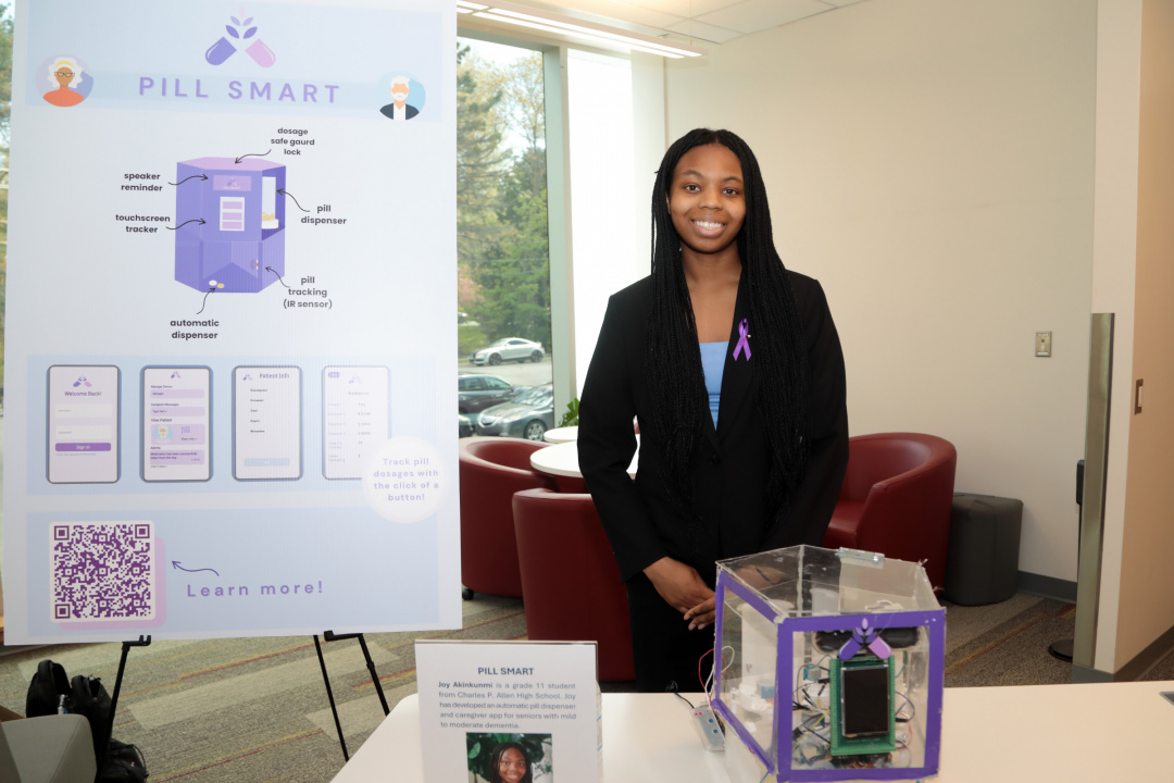 Student Joy Akinkunmi stands at a table with her Pill Smart device.