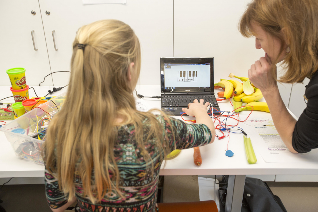 Une élève, assise à une table où se trouvent des bananes, des pots de Play-Doh et des fils raccordés à un ordinateur portable, utilise un projet de technologie interactive sous la supervision d’une personne enseignante.