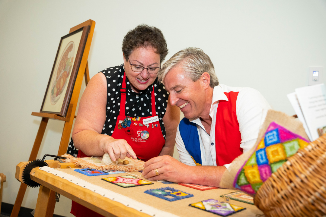 Premier Tim Houston gets a lesson in traditional Chéticamp rug hooking from local expert Betty Ann Cormier