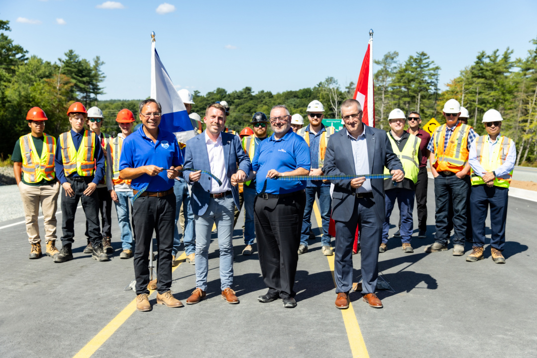 (left to right): Brian Wong, MLA for Waverley-Fall River-Beaver Bank, Braedon Clark, MP for Sackville-Bedford-Preston, Fred Tilley, Minister of Public Works, and Mark Underhill, General Manager of Highway Operations Nova Scotia with Dexter Construction, Vice President of Municipal Group cut the ribbon at the opening of the Aerotech Connector with road workers standing behind them.
