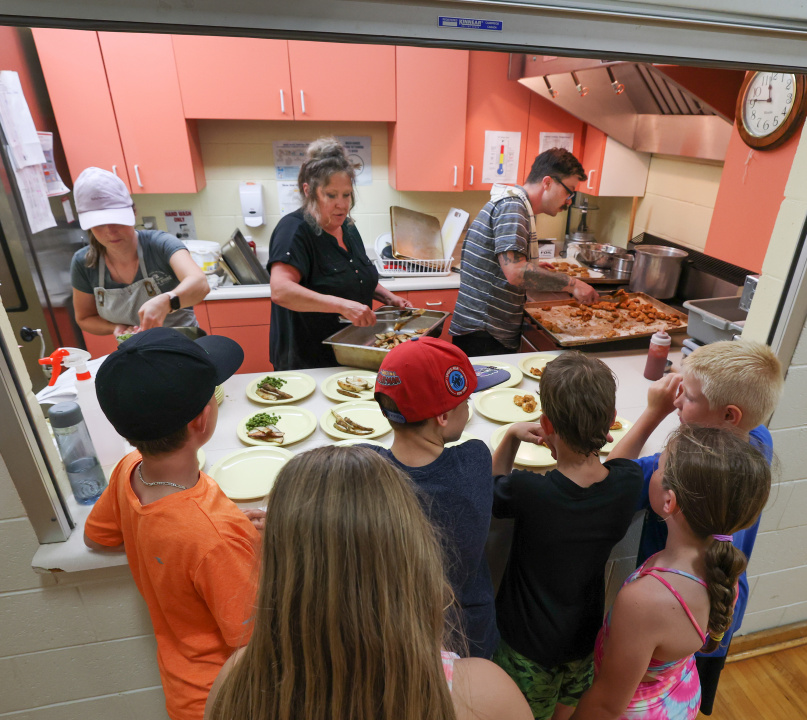 Students gather outside a school kitchen to pick up lunch as lunch program staff prepare meals.
