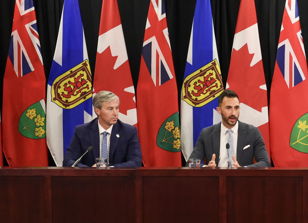 Premier Tim Houston and Ontario Minister Stephen Lecce at a desk in front of flags