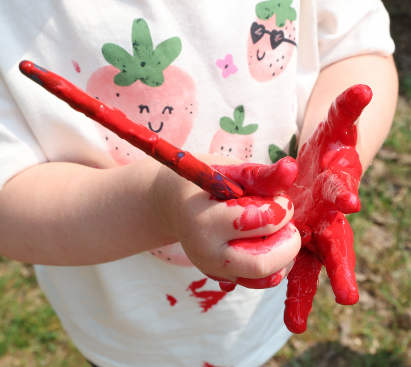 A youngster's hands holding a paint brush and covered in red paint.
