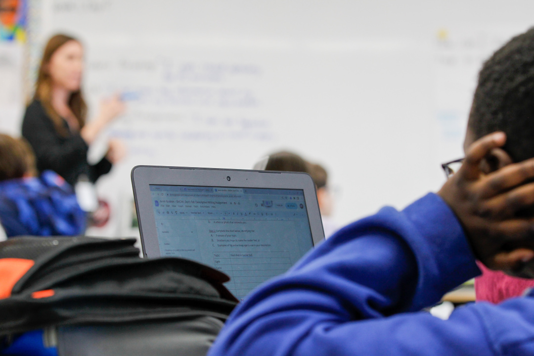 A student looks at a screen while listening to the teacher standing at the front of the classroom. 