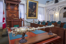 Photo showing mace sitting on a table in front of MLAs’ desks in the legislative chamber at Province House. The Speaker’s desk and a large portrait are in the background