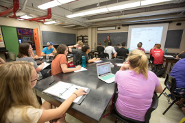 Students in a classroom sitting at tables looking towards a projector screen